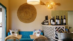 Un rincón acogedor con un sofá azul, dos sillas de mimbre y una mesa con un menú. Se ve un área de bar con botellas de vino y una luz decorativa al fondo.