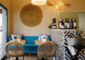 Un'accogliente area salotto con un divano blu, due poltrone in vimini e un tavolo con il menu. Sullo sfondo, si intravede una zona bar con bottiglie di vino e una luce decorativa.