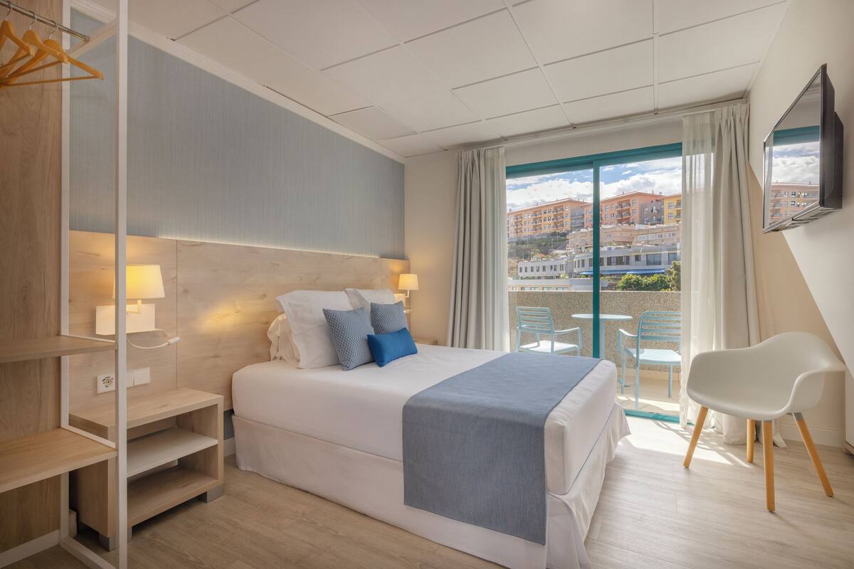 A hotel room with a bed, bedside tables, a TV, a balcony with a view of buildings.