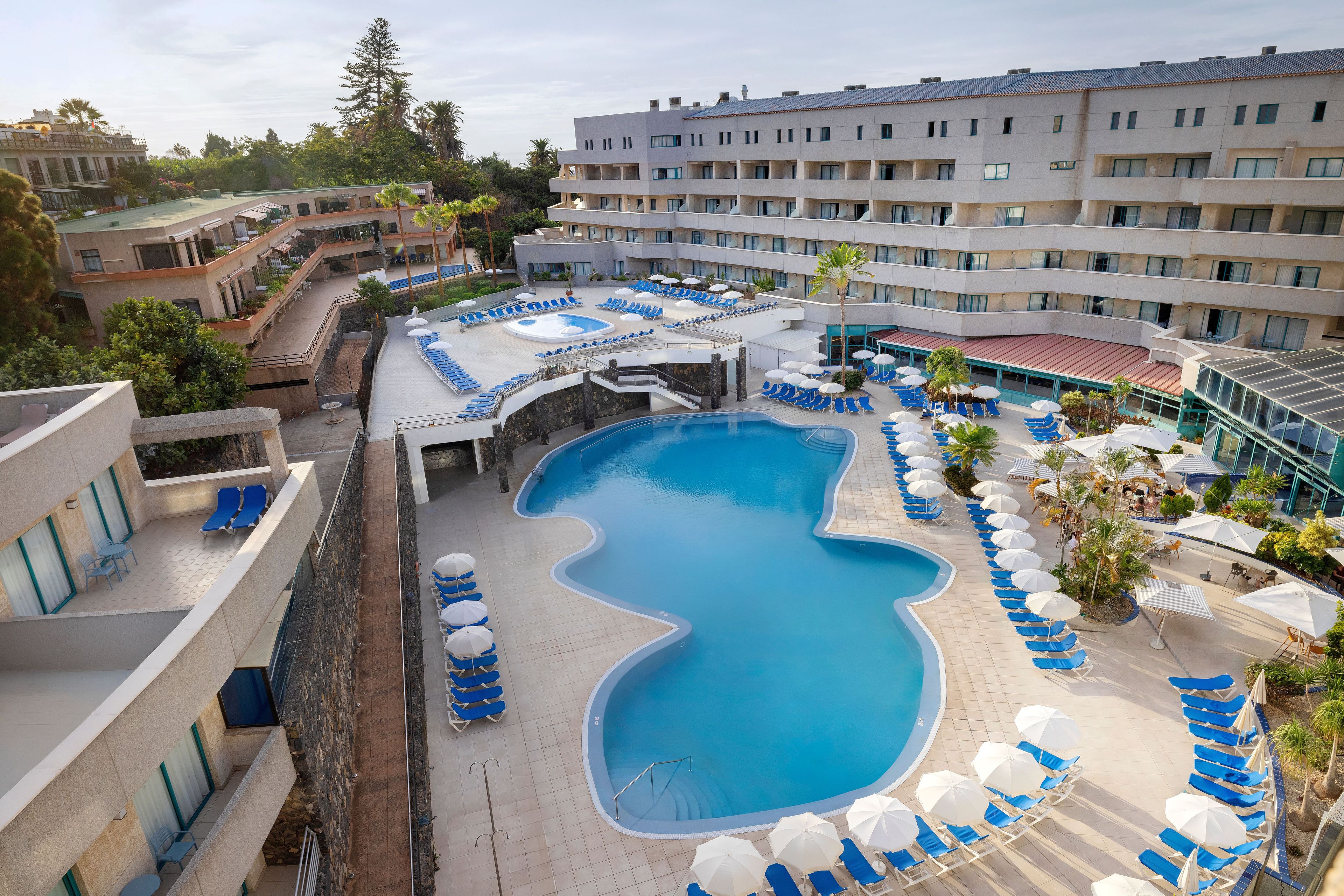 Ein Hotel mit einem großen Schwimmbecken, umgeben von Liegestühlen und Sonnenschirmen, mehreren Etagen sowie einer Mischung aus moderner und traditioneller Architektur.