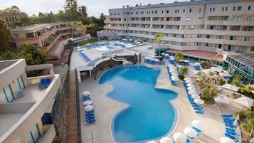 Un hotel con una amplia piscina rodeada de tumbonas y sombrillas, varios pisos y una combinación de arquitectura moderna y tradicional.