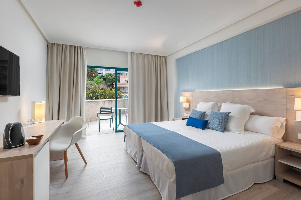 A hotel room with a bed, bedside table, lamp, desk, chair, and a view of the outdoors.