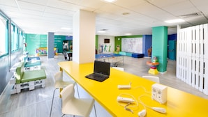 Un área de espera luminosa y moderna, con un mostrador amarillo, sillas blancas y una zona de juegos verde.