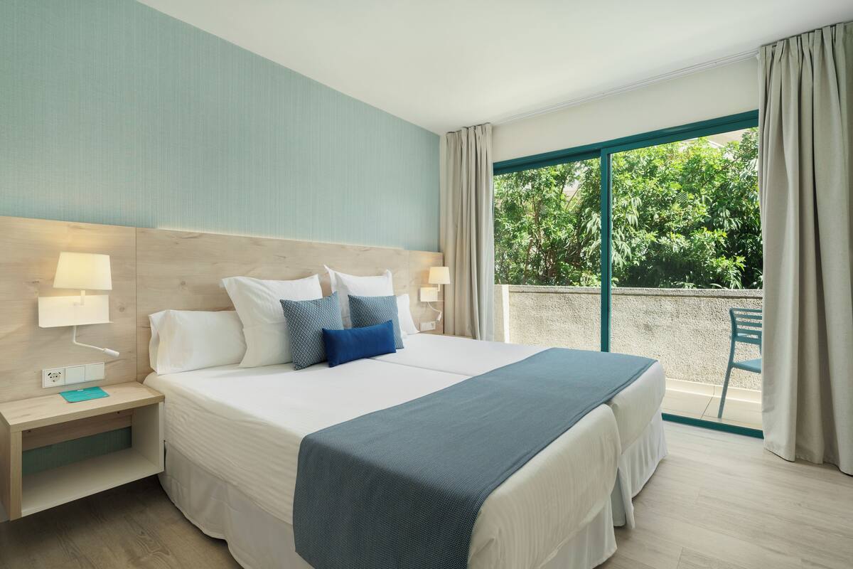 A hotel room with a large bed, two bedside tables with lamps, a view of greenery through a sliding glass door, and a wooden floor.