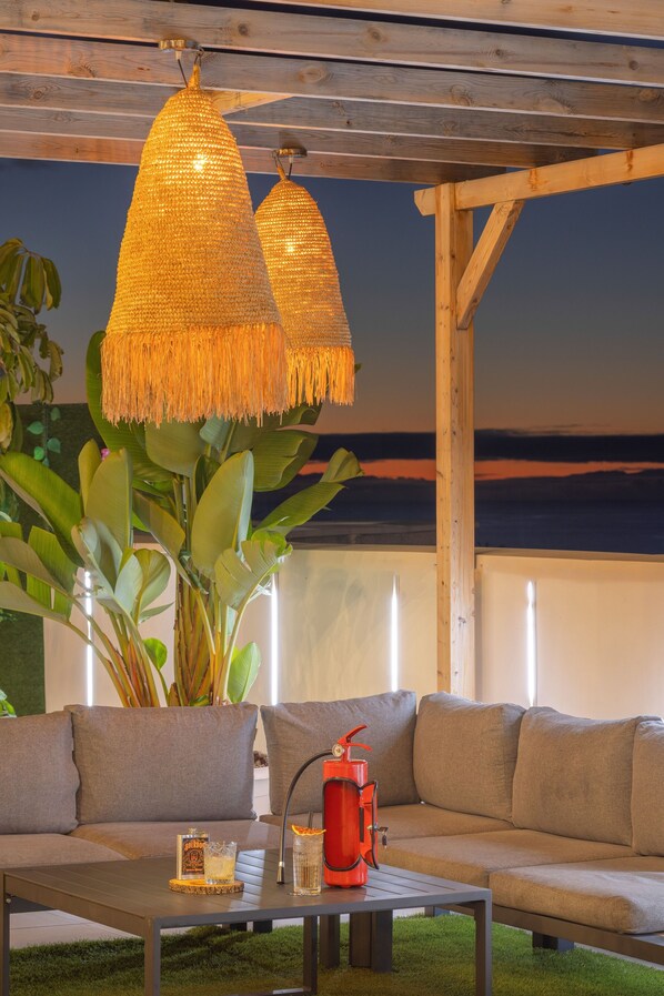 Un'accogliente area salotto esterna con una pergola in legno, un divano e un tavolo con bevande e un contenitore rosso.
