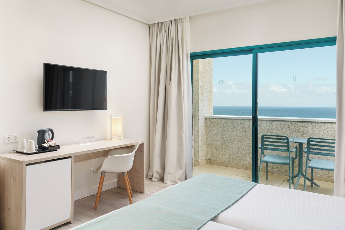 Camera d'albergo con letto, scrivania, sedia, televisione e un balcone con vista sul mare.