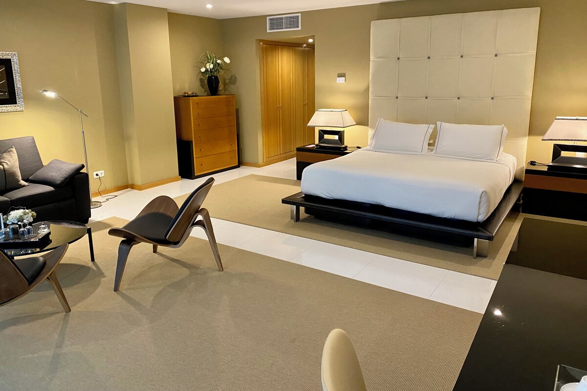 A modern hotel room with a large bed, a sofa, a chair, a desk, and a nightstand with lamps.