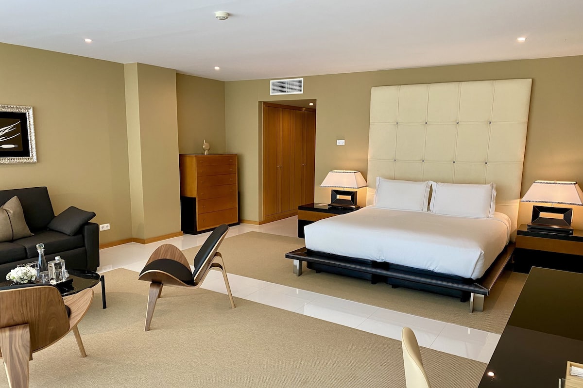 A hotel room with a large bed, two bedside tables with lamps, a wooden dresser, a black sofa, a small table with decorative items, and a chair.