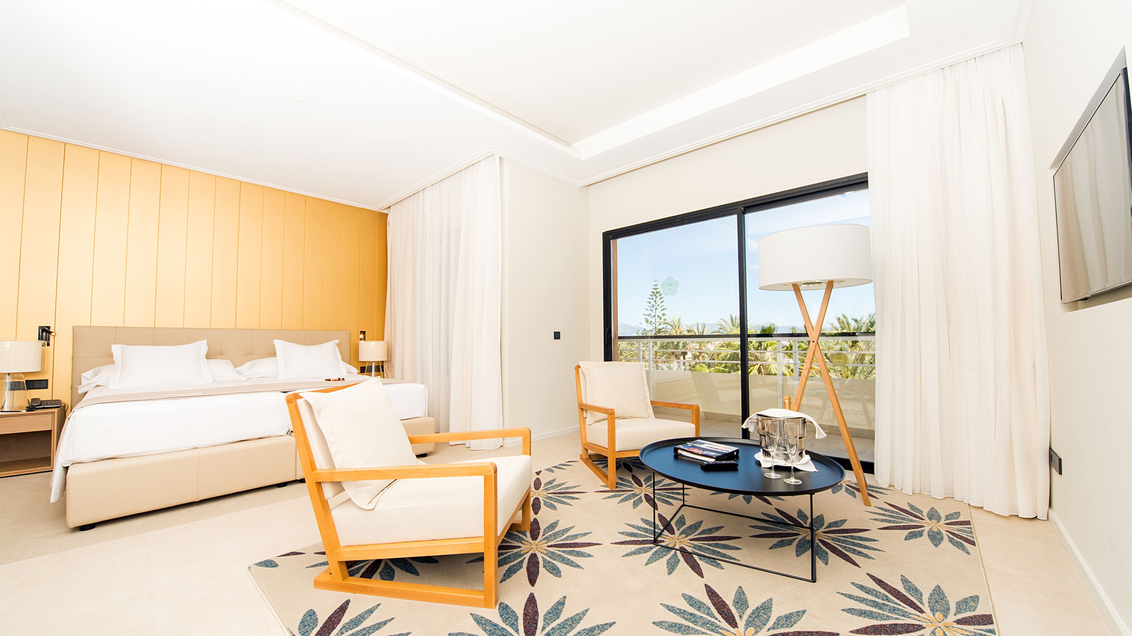 Habitación de hotel con cama, dos sillas, una mesita con lámpara y un ventanal con vistas.
