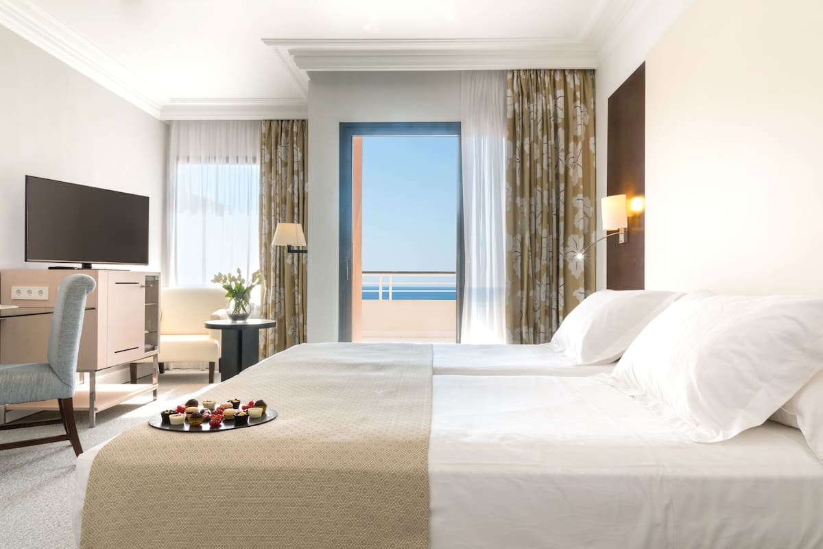 A hotel room with a bed, a desk with a TV, a chair, a small table with a lamp, and a balcony with curtains.