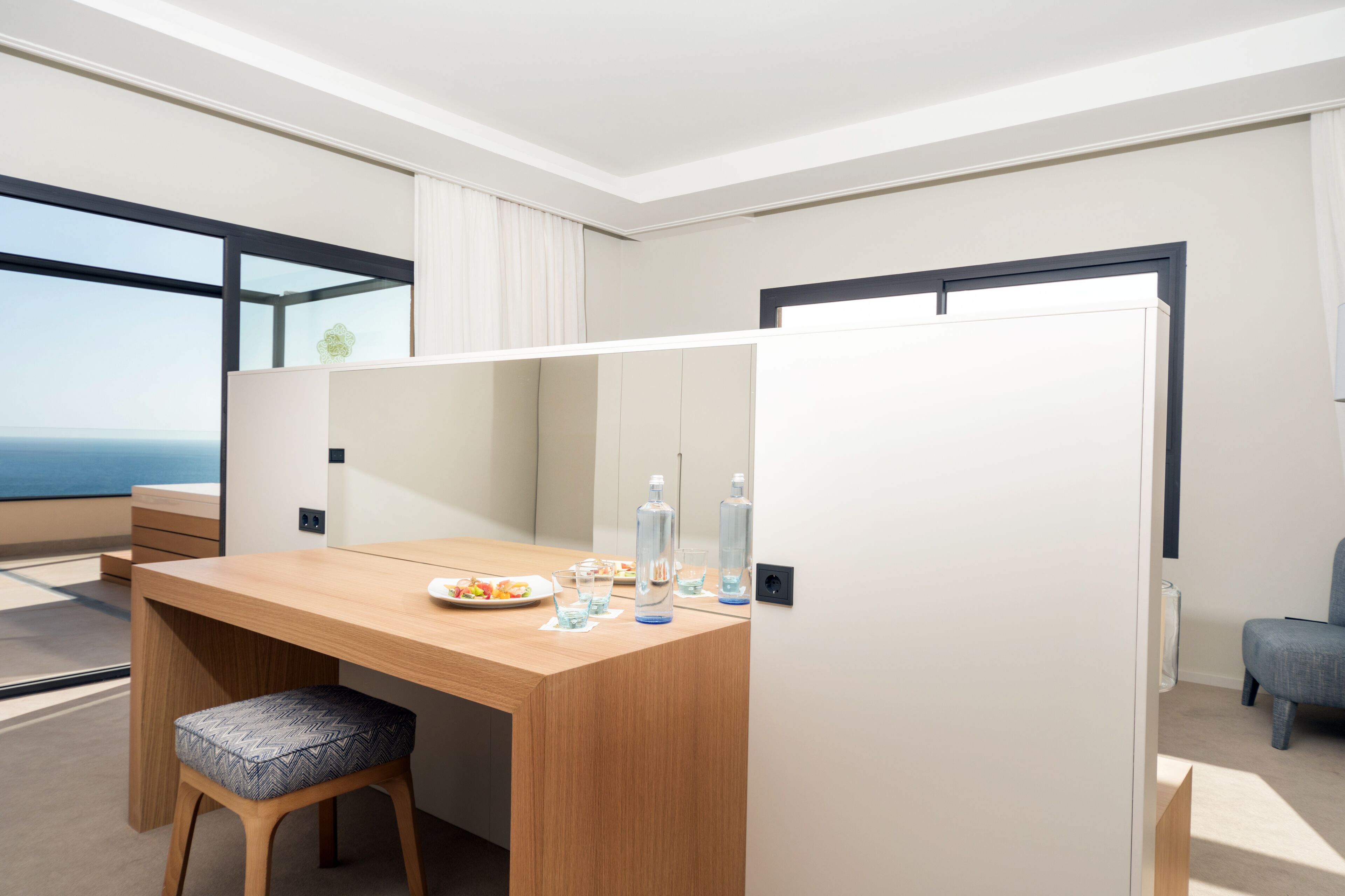 A modern hotel room with a wooden desk, a cushioned stool, and a large window offering a view of the sea.
