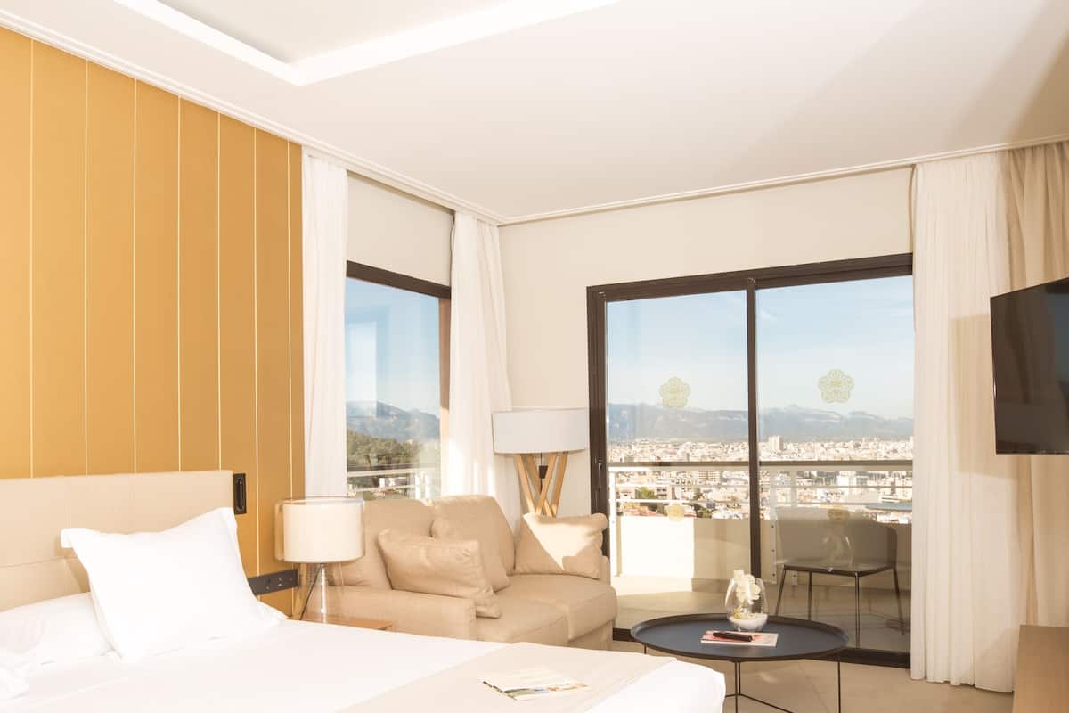 A hotel room with a bed, a sofa, a TV, a balcony with a city view, and a table with a vase of flowers.