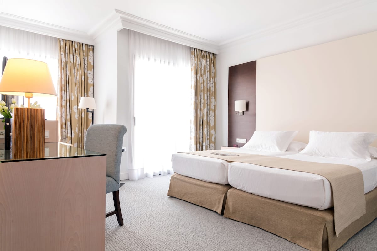A hotel room with a bed, a desk, a chair, a lamp, and curtains.