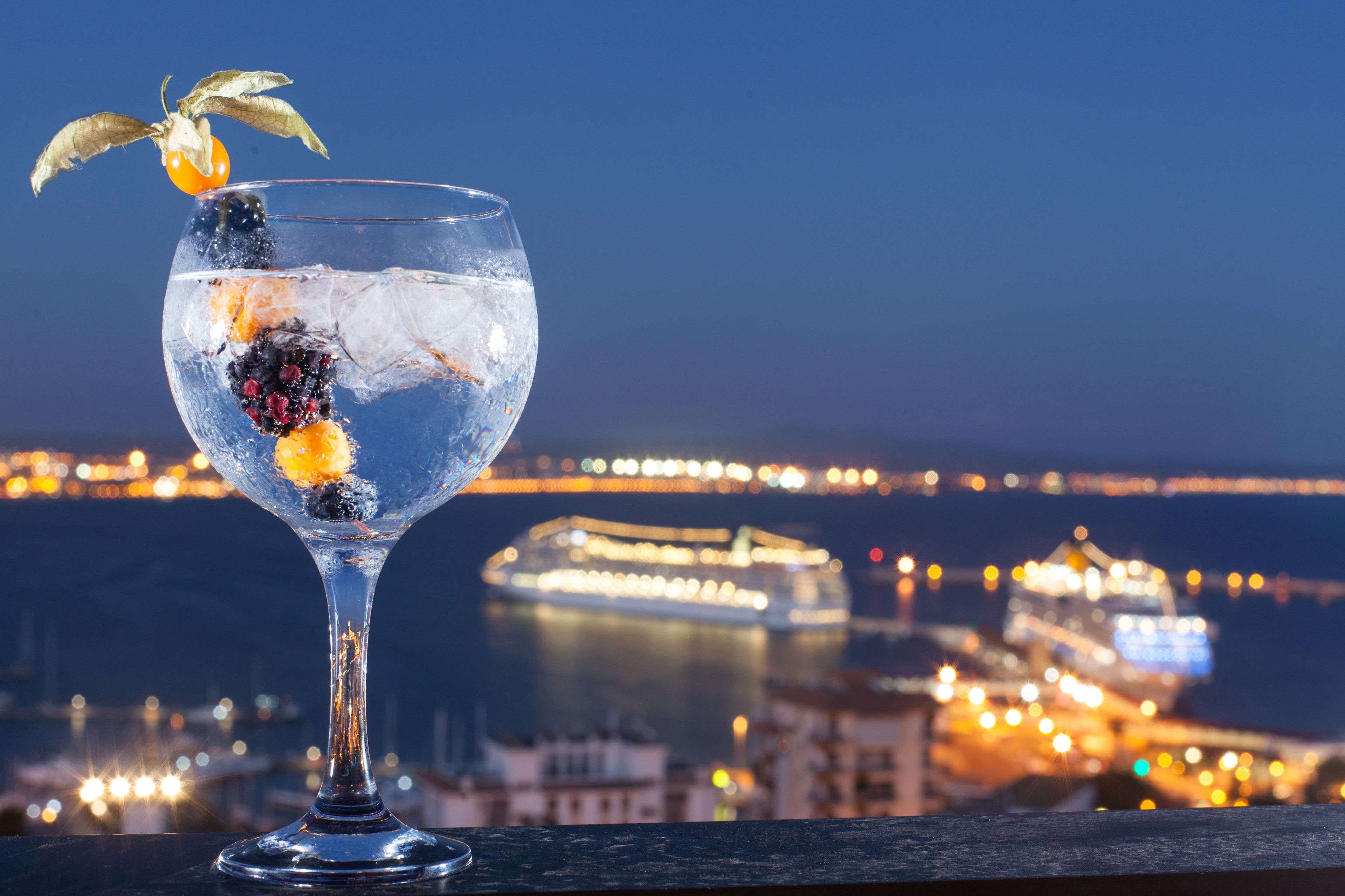 Ein Cocktailglas mit Fruchtdekoration auf einem Balkon mit Blick auf eine nächtliche Stadtlandschaft.