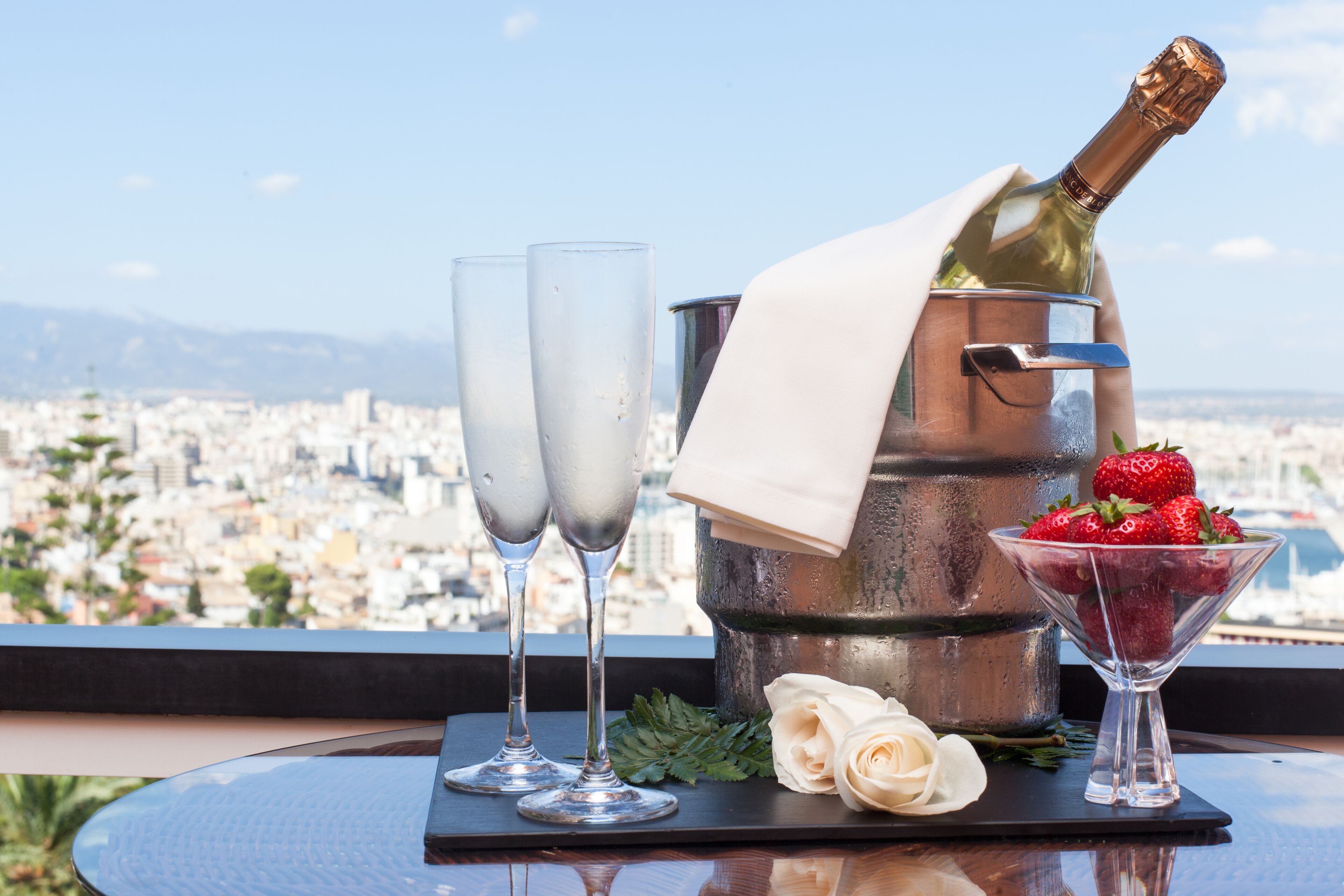 Una mesa con dos copas de champagne, una botella de champagne, una copa de martini con fresas y una rosa blanca en un balcón con vistas a un paisaje urbano.