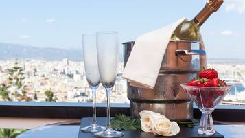 Una mesa con dos copas de champagne, una botella de champagne, una copa de martini con fresas y una rosa blanca en un balcón con vistas a un paisaje urbano.