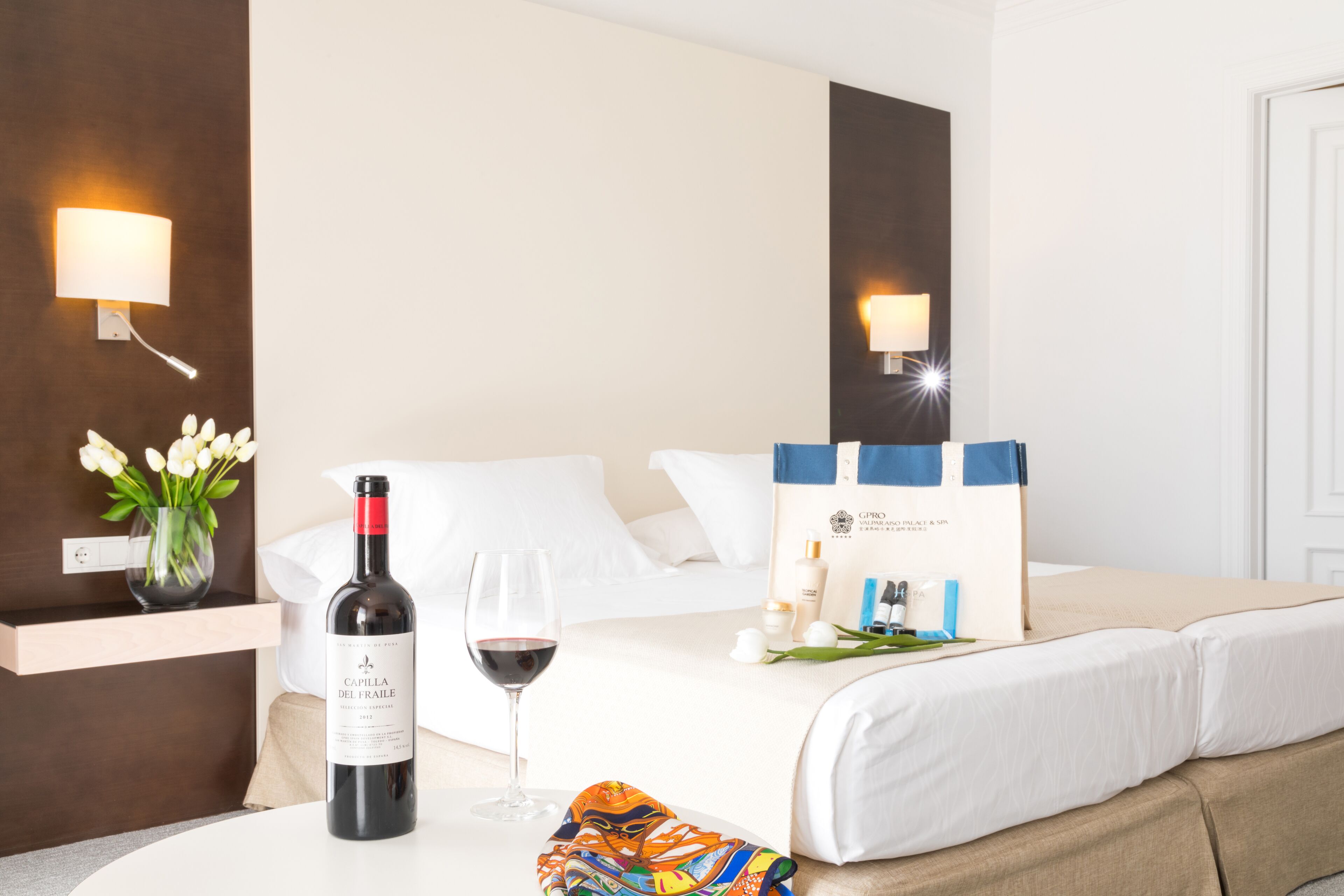 Habitación de hotel con una cama, una botella de vino, una copa de vino, un jarrón con flores y un neceser de artículos de aseo.