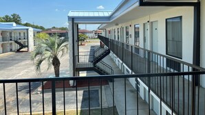 Corredor de hotel com área externa coberta, uma palmeira e um estacionamento ao fundo.
