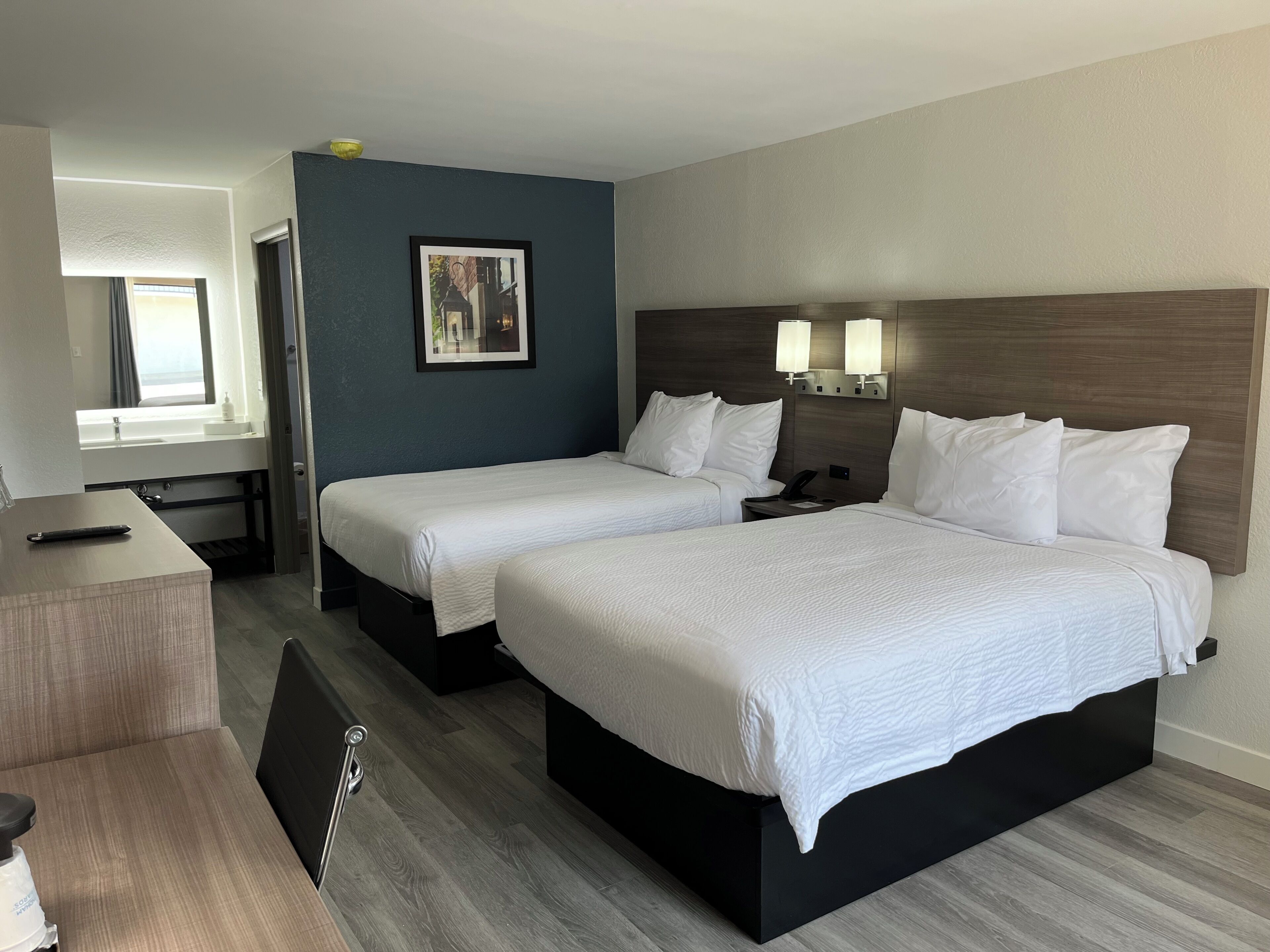 A hotel room with two beds, a desk, a chair, a nightstand, and a wall-mounted lamp.