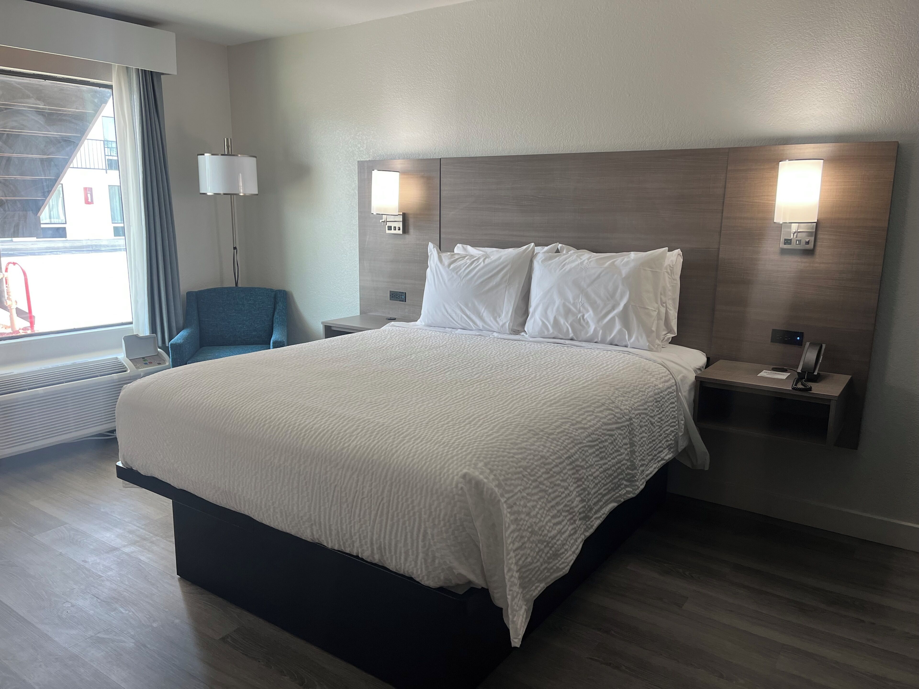 A hotel room with a large bed, bedside tables, a desk, a chair, a window with curtains, and wall-mounted lamps.