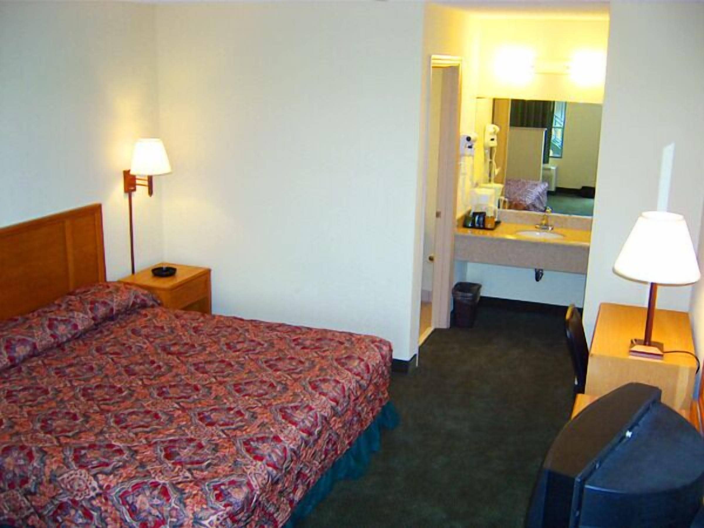 A hotel room with a bed, a desk, a chair, a mirror, and a sink.