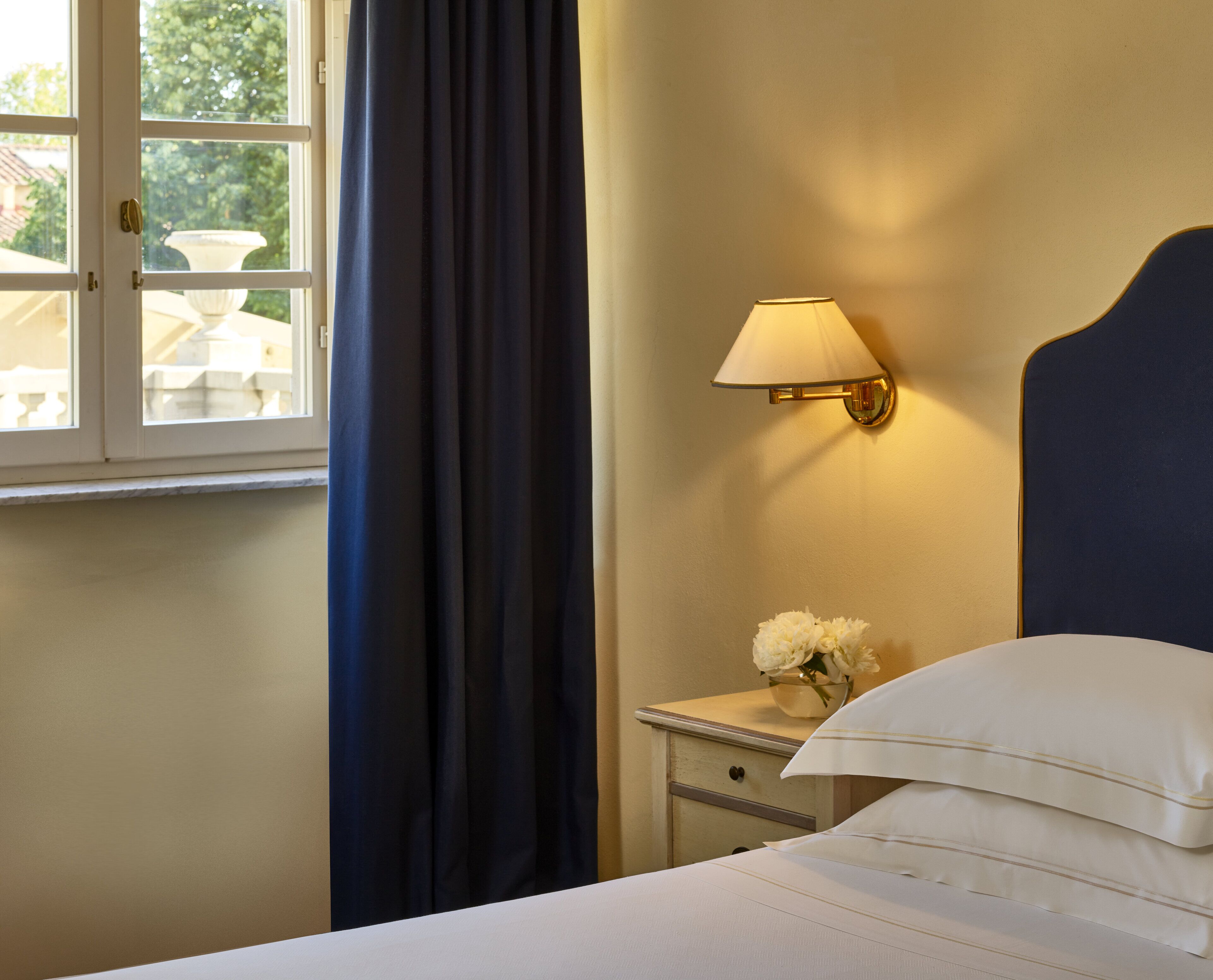 Een hotelkamer met een bed, nachtkastje met lamp, vaas met bloemen en een raam met gordijnen.