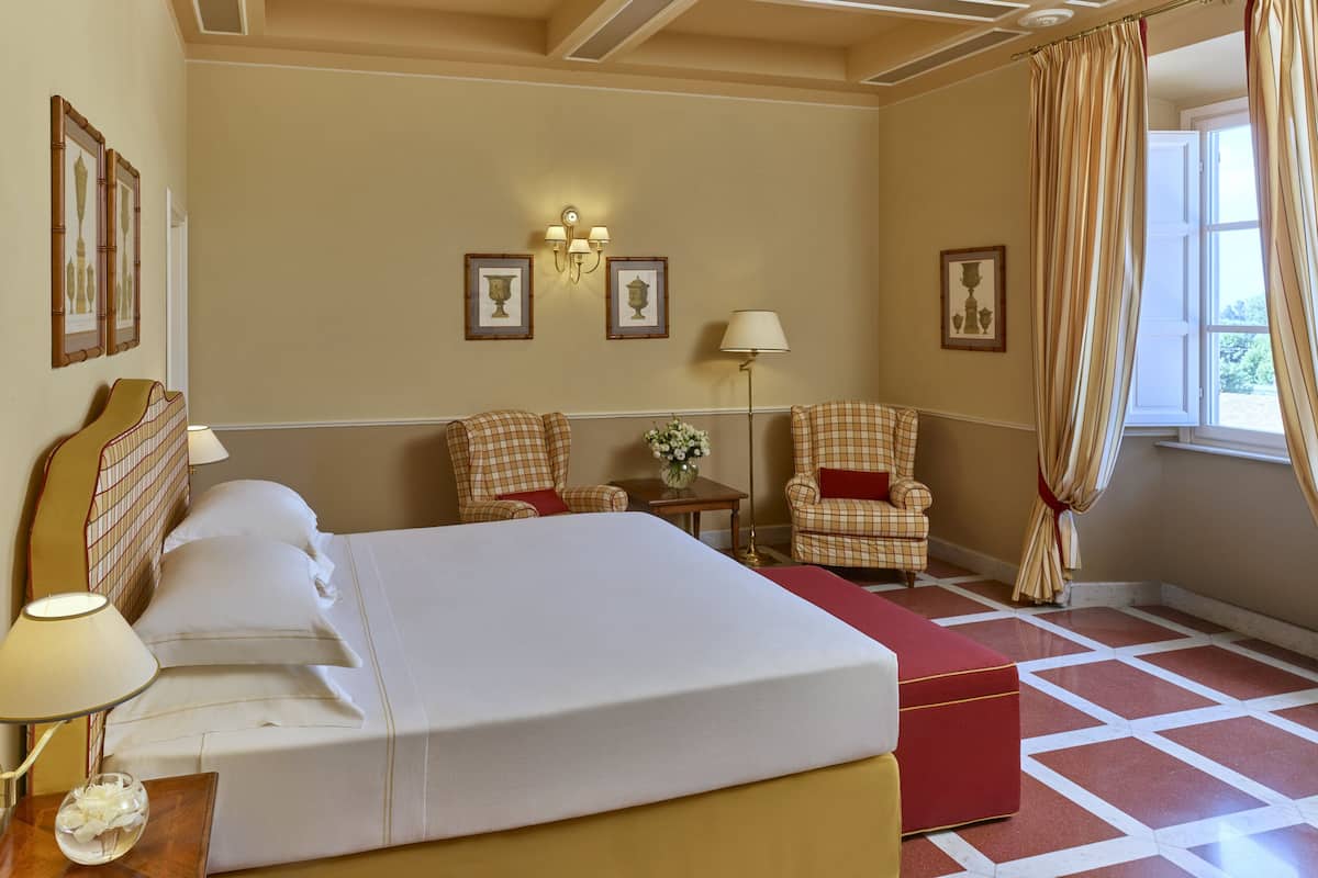 A hotel room with a large bed, two armchairs, a small table with a vase, and a window with curtains.