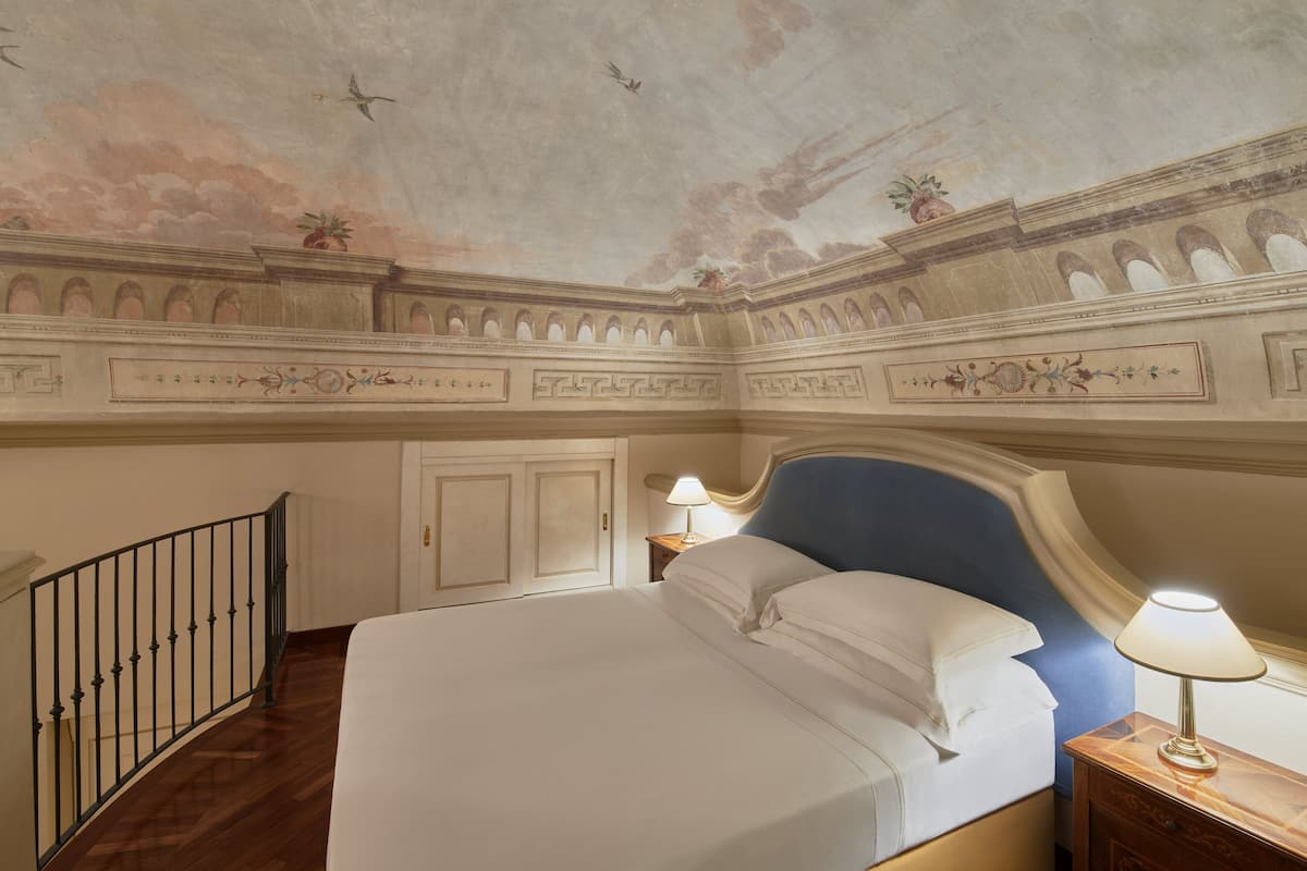 A hotel room with a large bed, a nightstand with a lamp, a ceiling with decorative frescoes, and a balcony with a railing.