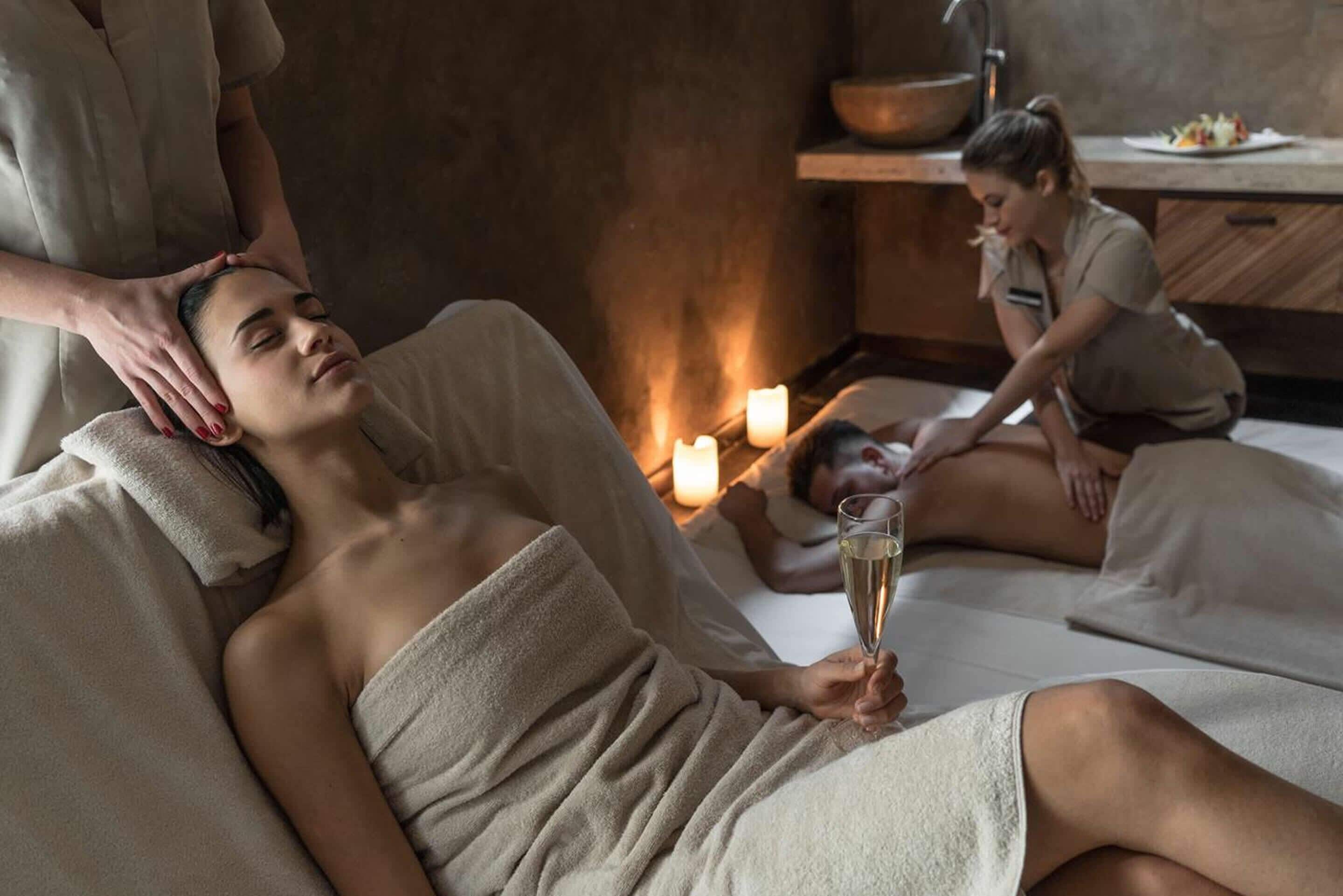 Un'atmosfera rilassante in una spa, con una persona che riceve un massaggio alla testa, un'altra seduta con un drink e luci di candela.