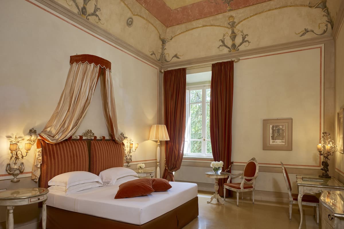 A classic four-poster bed with a canopy, a sitting area with a chair and table, and a large window with curtains.