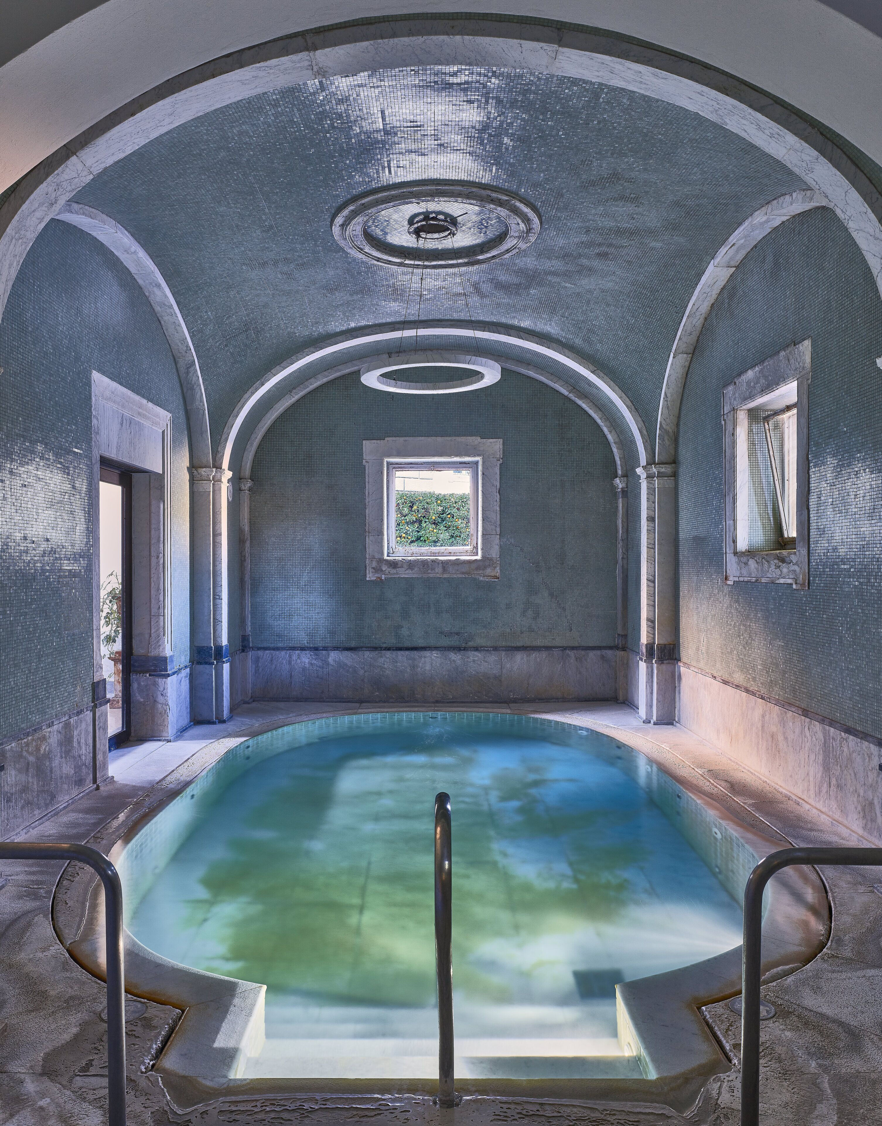 An indoor swimming pool with a clear view of the ceiling and walls, featuring a circular skylight and a small window.