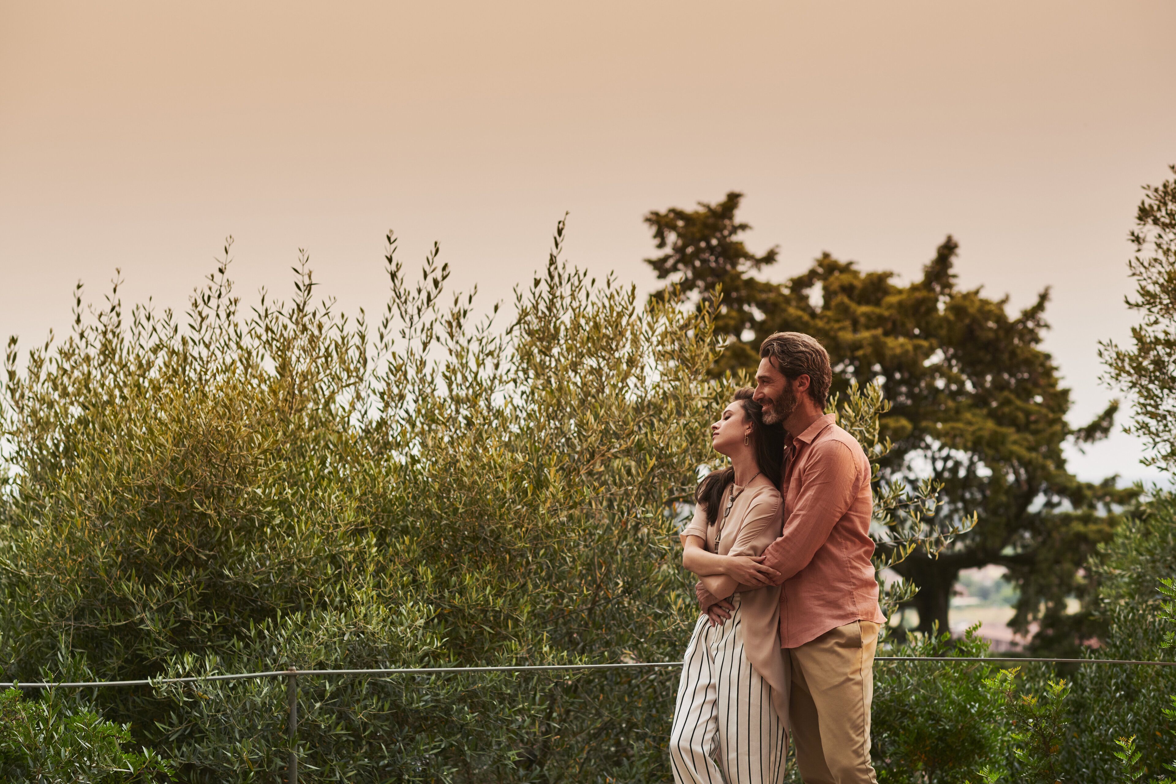 A couple embracing in a natural setting with dense foliage and trees in the background.