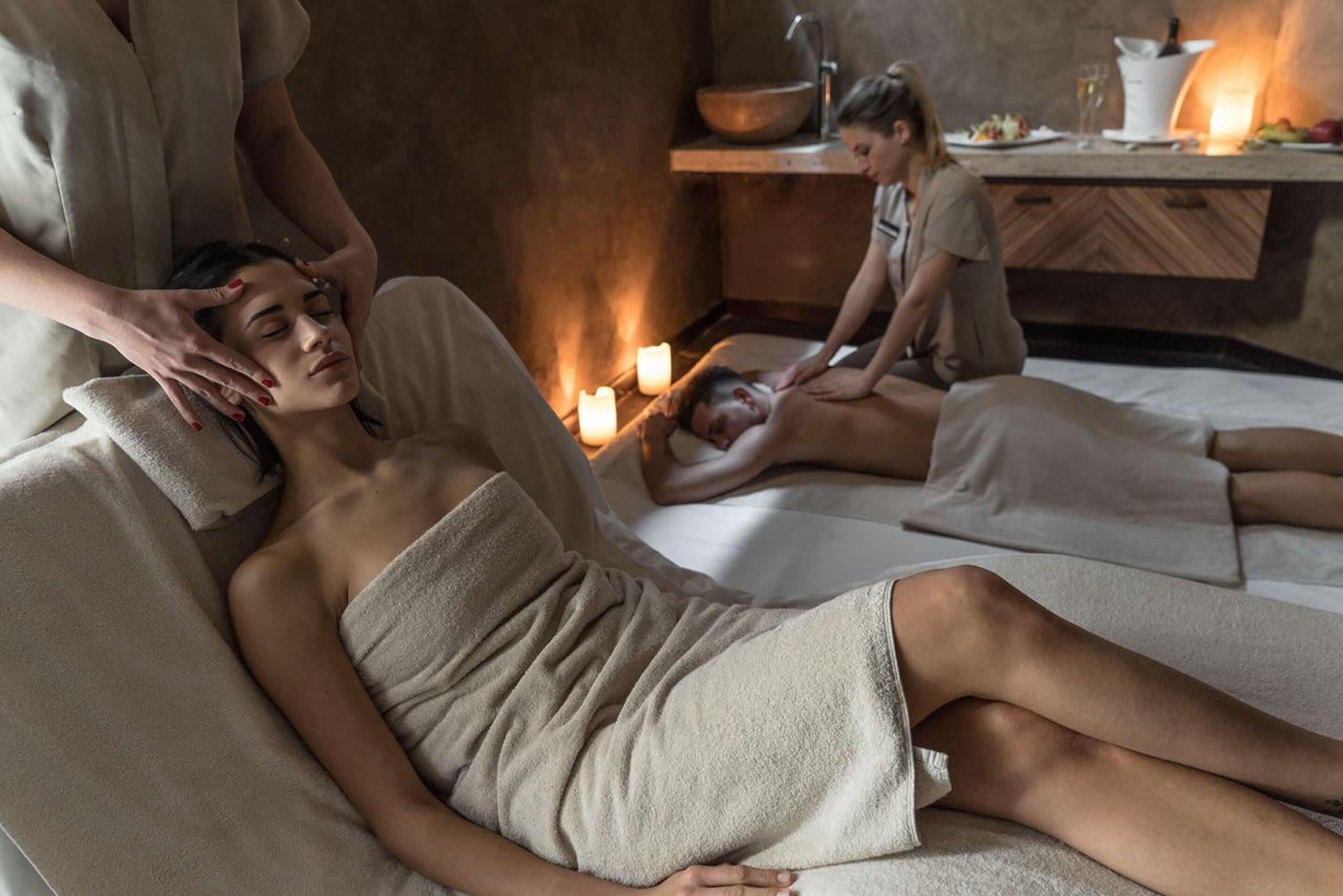 Ein Wellnessraum mit zwei Massagetischen, Kerzen und einer Holztresse mit Schale und Glas.
