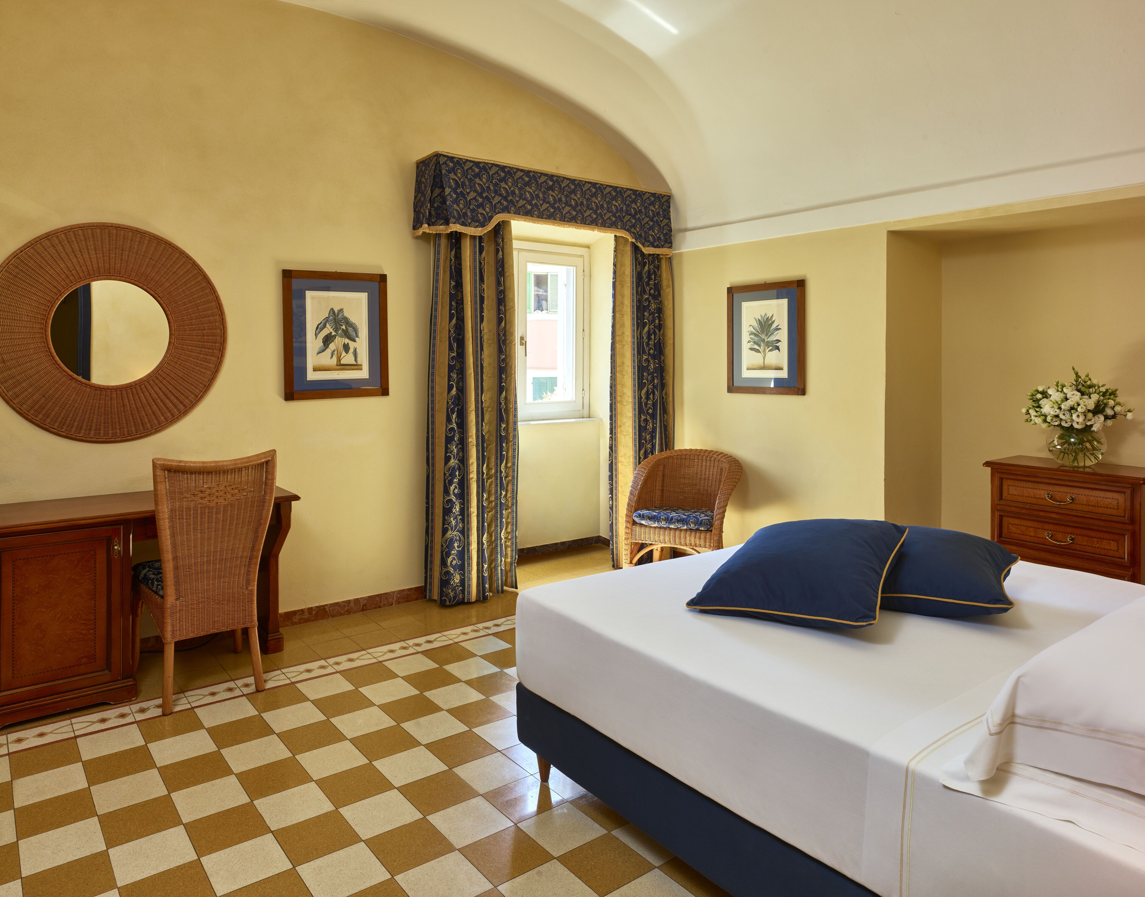 A hotel room with a checkered floor, a bed with blue pillows, a wicker chair, a wooden dresser, and two framed pictures on the wall.