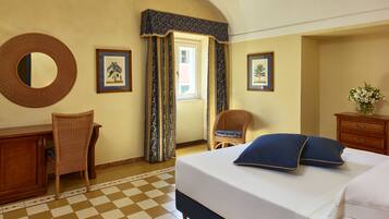 A hotel room with a checkered floor, a bed with blue pillows, a wicker chair, a wooden dresser, and two framed pictures on the wall.
