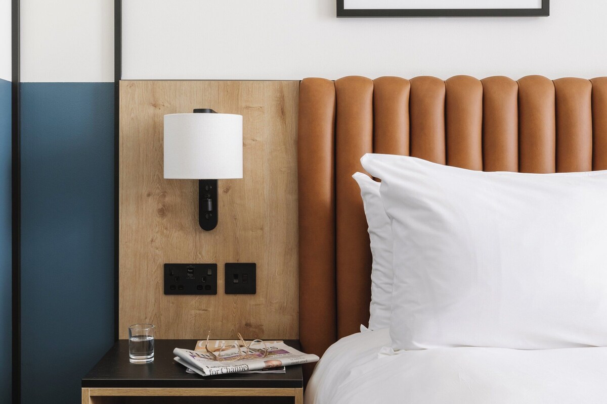 Een hotelkamer met een houten hoofdeinde, een nachtkastje met een lamp, een tijdschrift en een glas water.