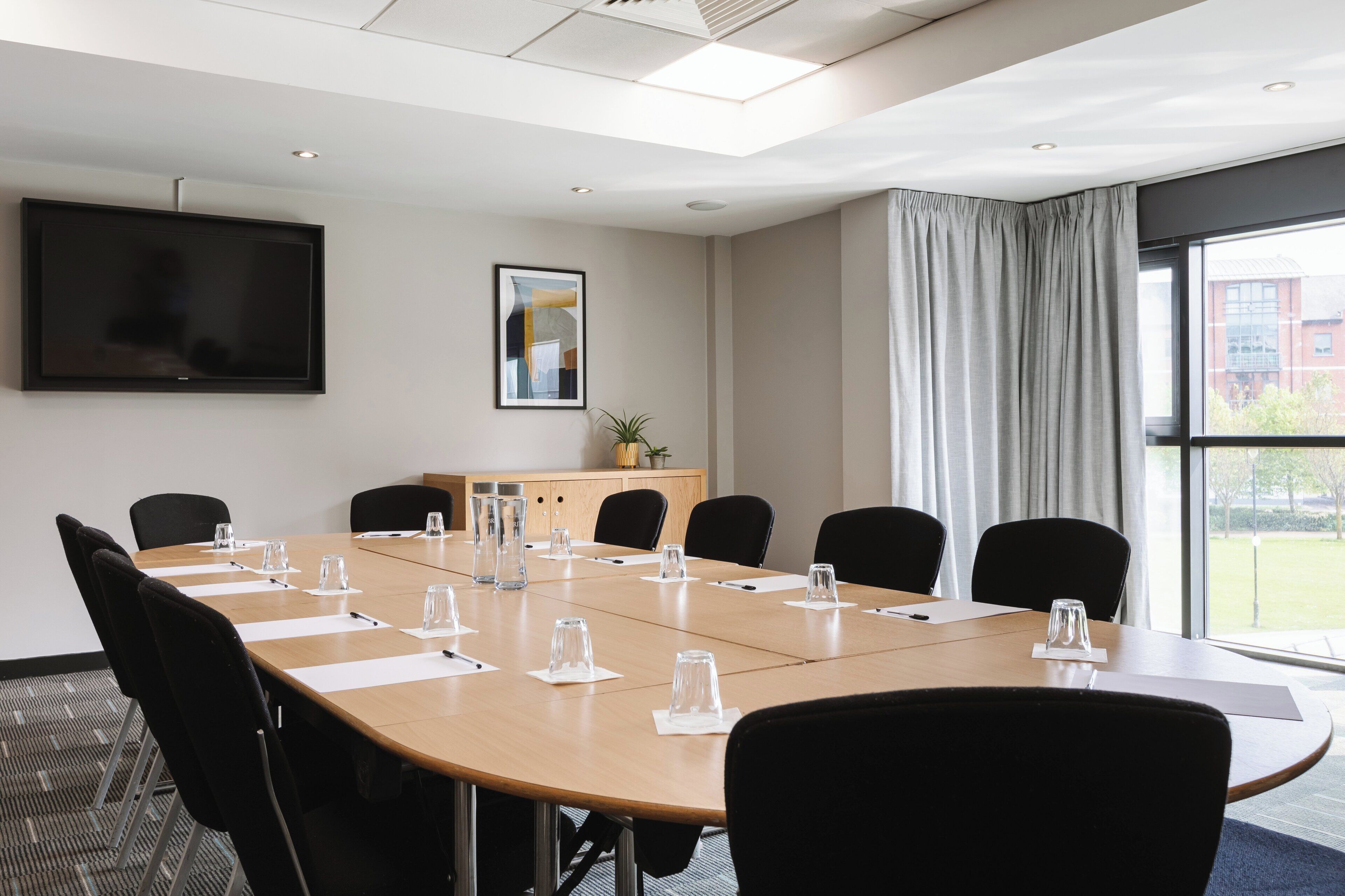A conference room with a long table, black chairs, a flat-screen TV, a framed picture, a cabinet with a plant, and a window with curtains.