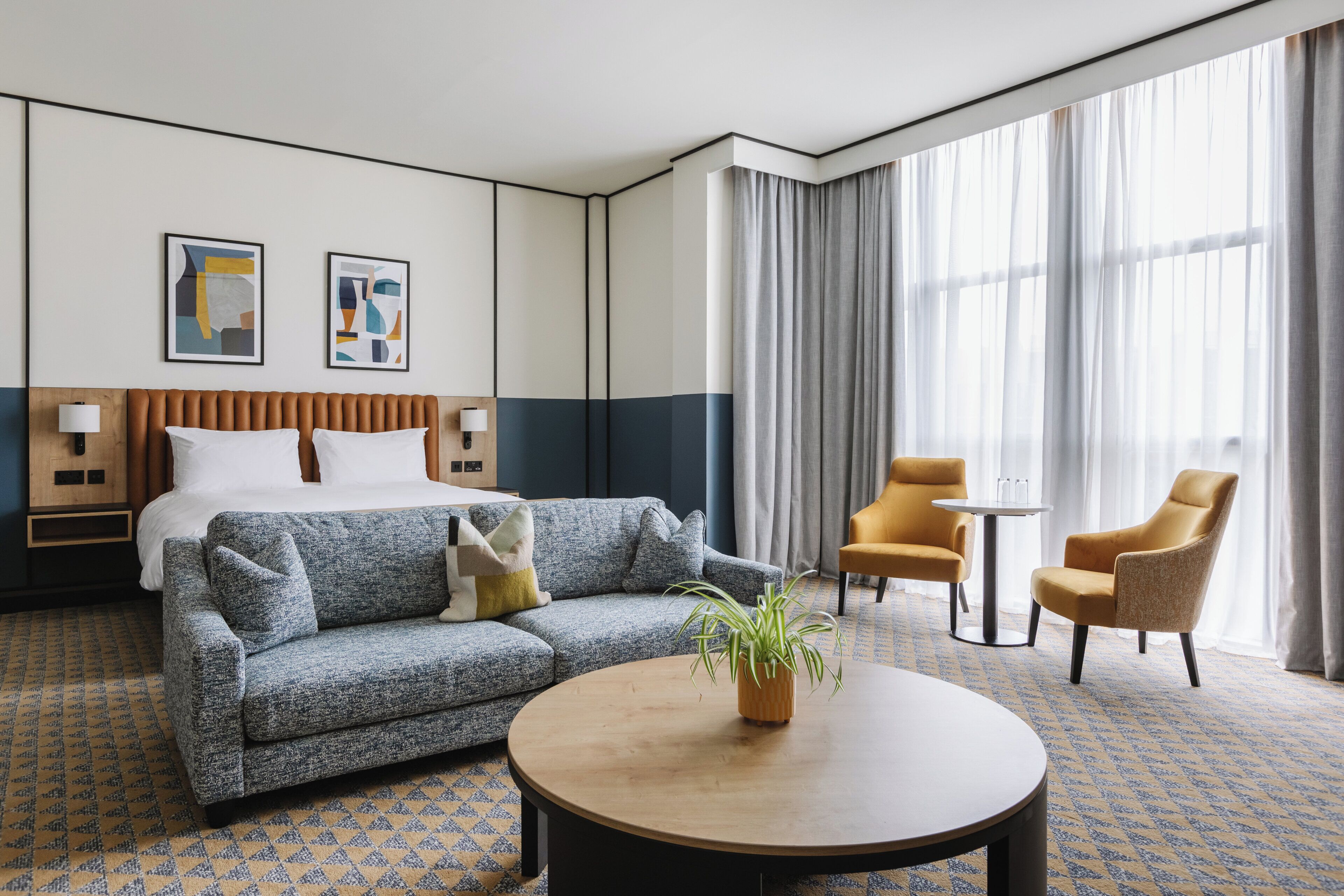 Habitación de hotel con cama, sofá, dos sillones, una mesita de centro, una planta y una ventana con cortinas.