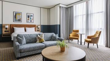Habitación de hotel con cama, sofá, dos sillones, una mesita de centro, una planta y una ventana con cortinas.
