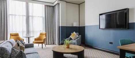 A modern hotel room with a sofa set, a round coffee table, a TV mounted on the wall, and a patterned carpet.