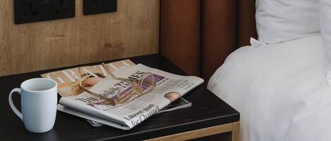 Habitación de hotel con cabecera de madera, mesita de noche con lámpara, una revista y una taza.