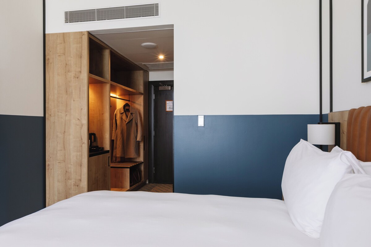 Een hotelkamer met een bed, een houten garderobe, een bureau en een nachtkastje met een lamp.