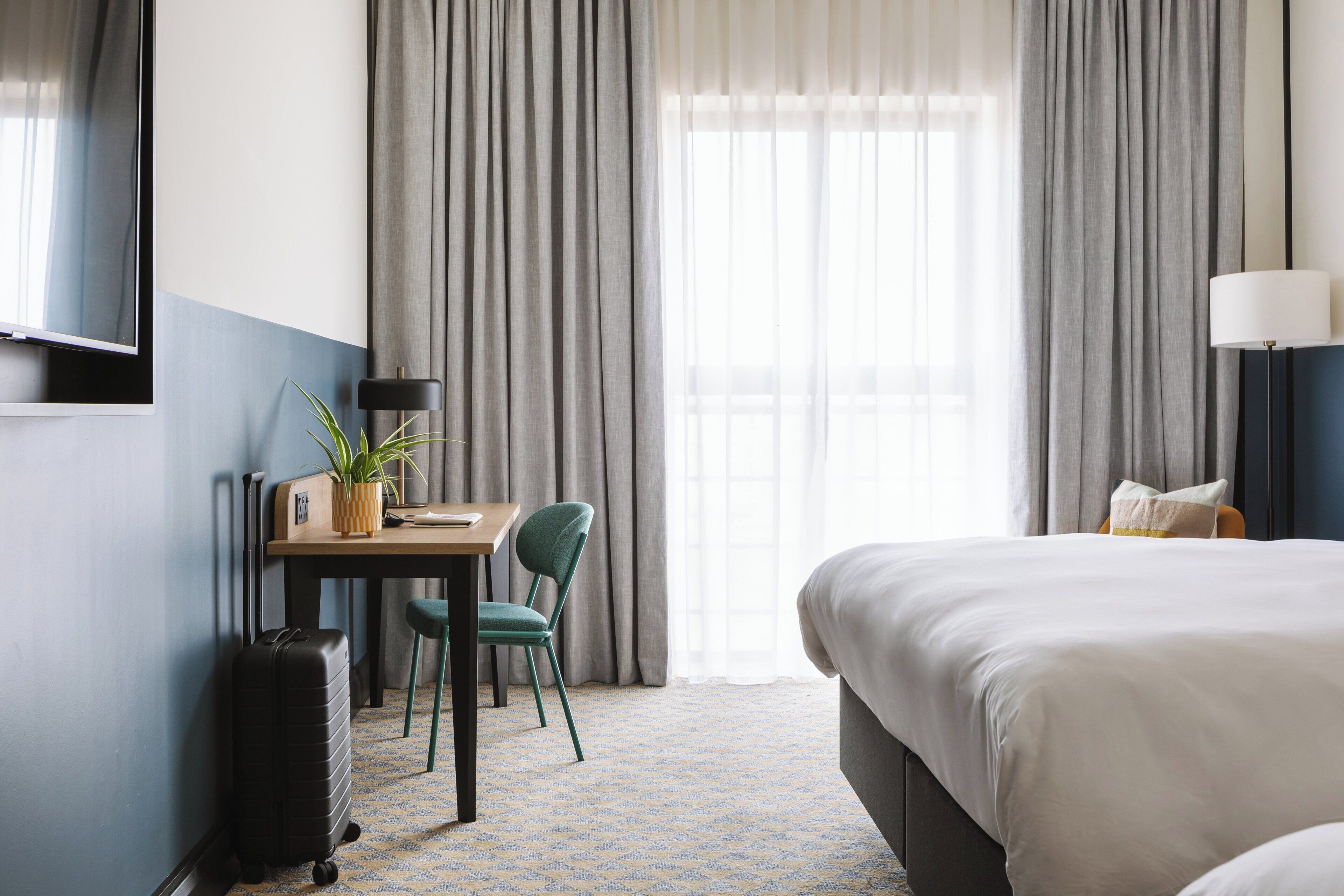 A hotel room with a bed, a desk with a chair, a lamp, a suitcase, and a window with curtains.