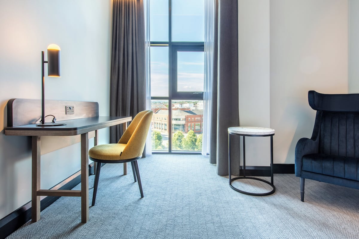 A hotel room with a desk, chair, and sofa, a large window with a view of buildings, and a small side table.