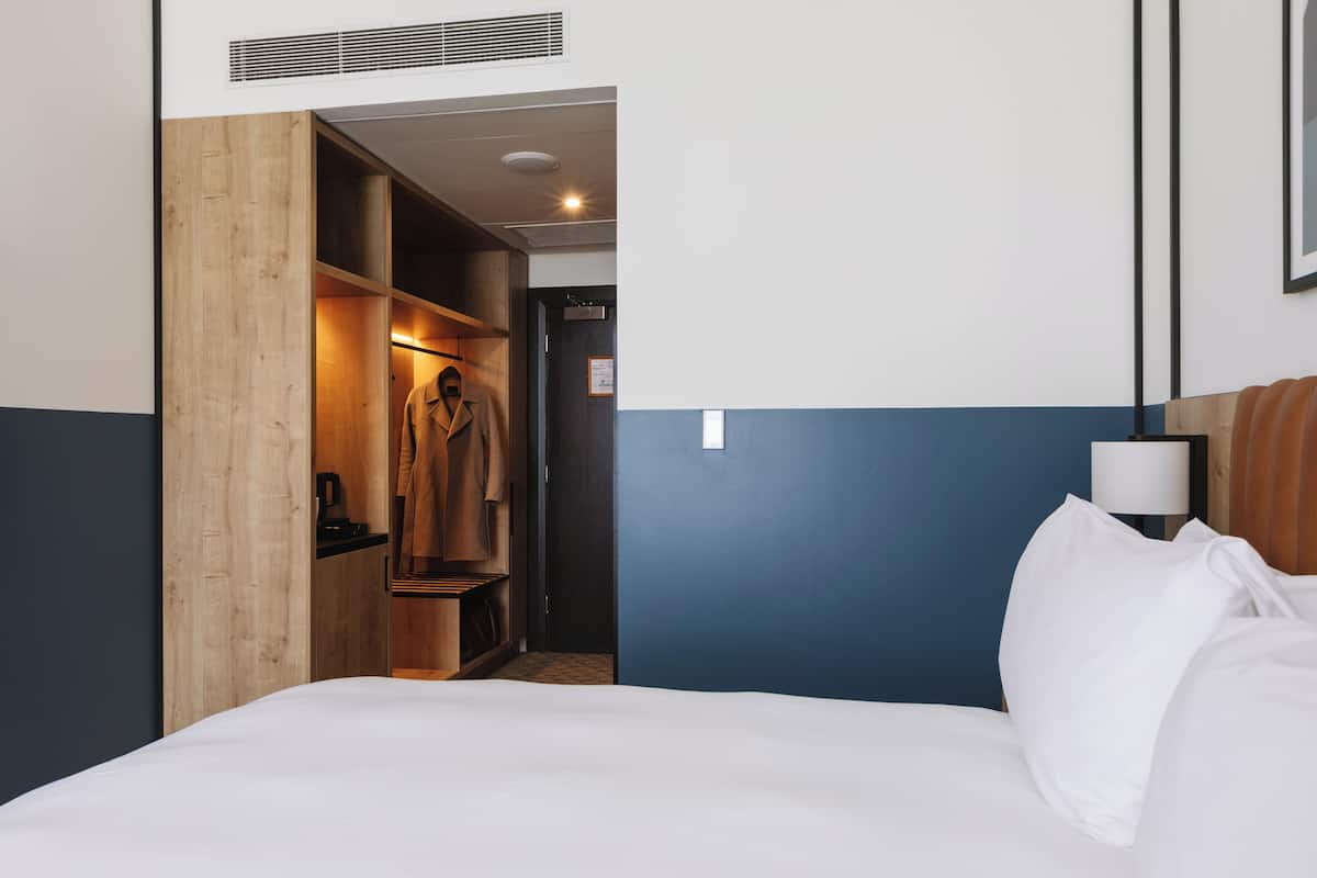 A hotel room with a bed, a wooden wardrobe, a desk, and a nightstand with a lamp.