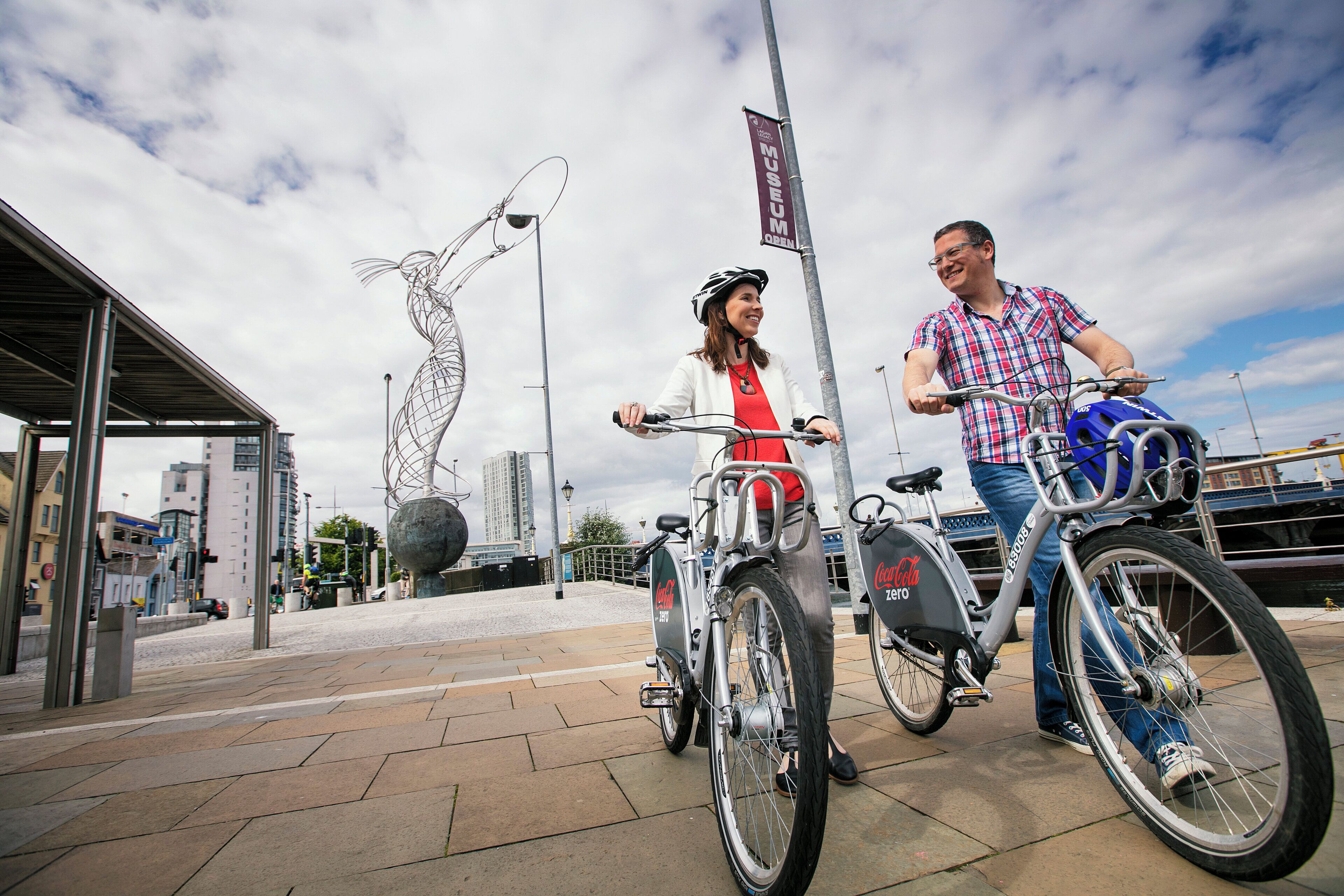 Two bicycles parked on a paved area with a metal sculpture in the background.