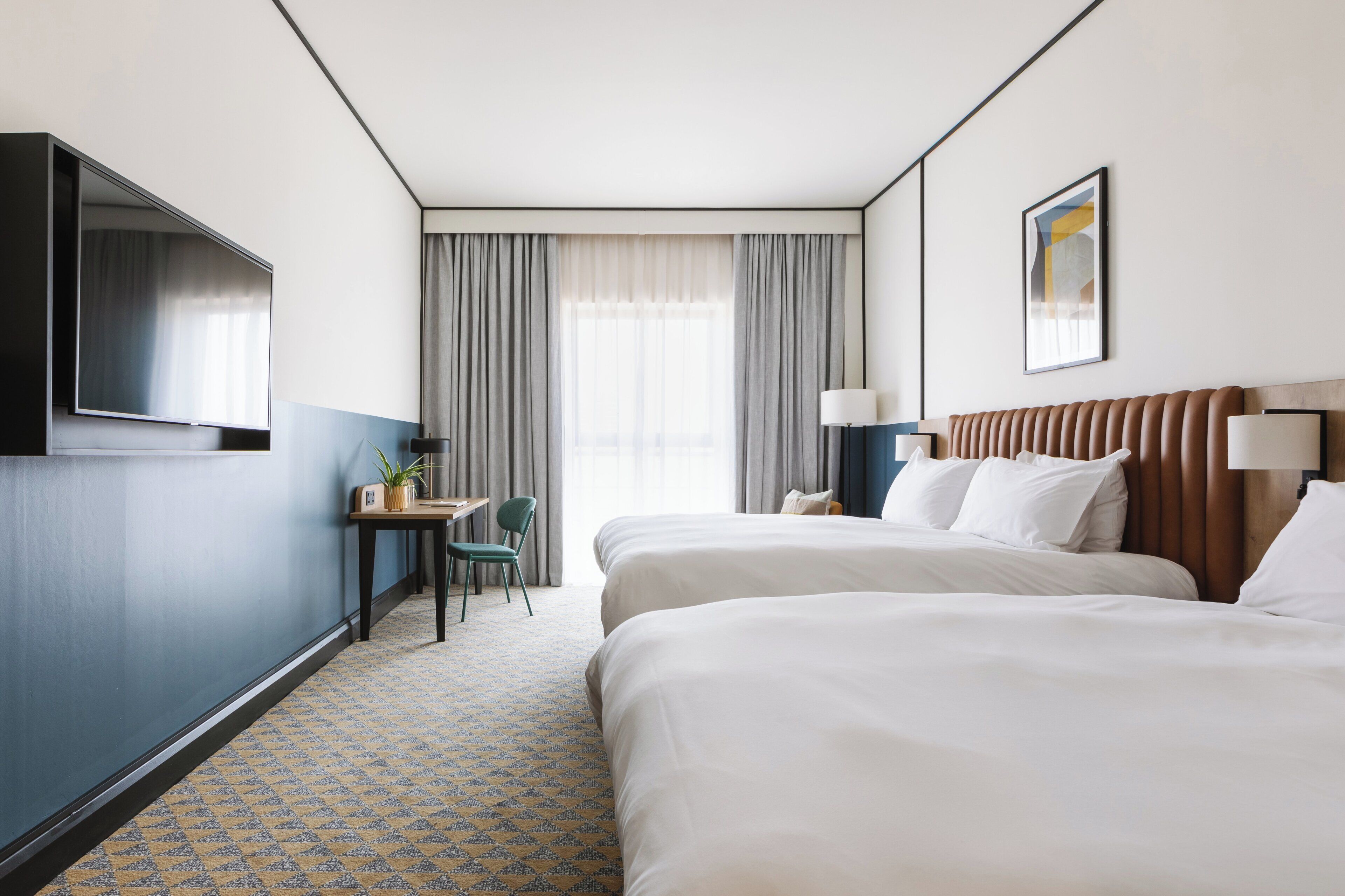 A hotel room with two beds, a desk, a chair, a TV, and a window with curtains.