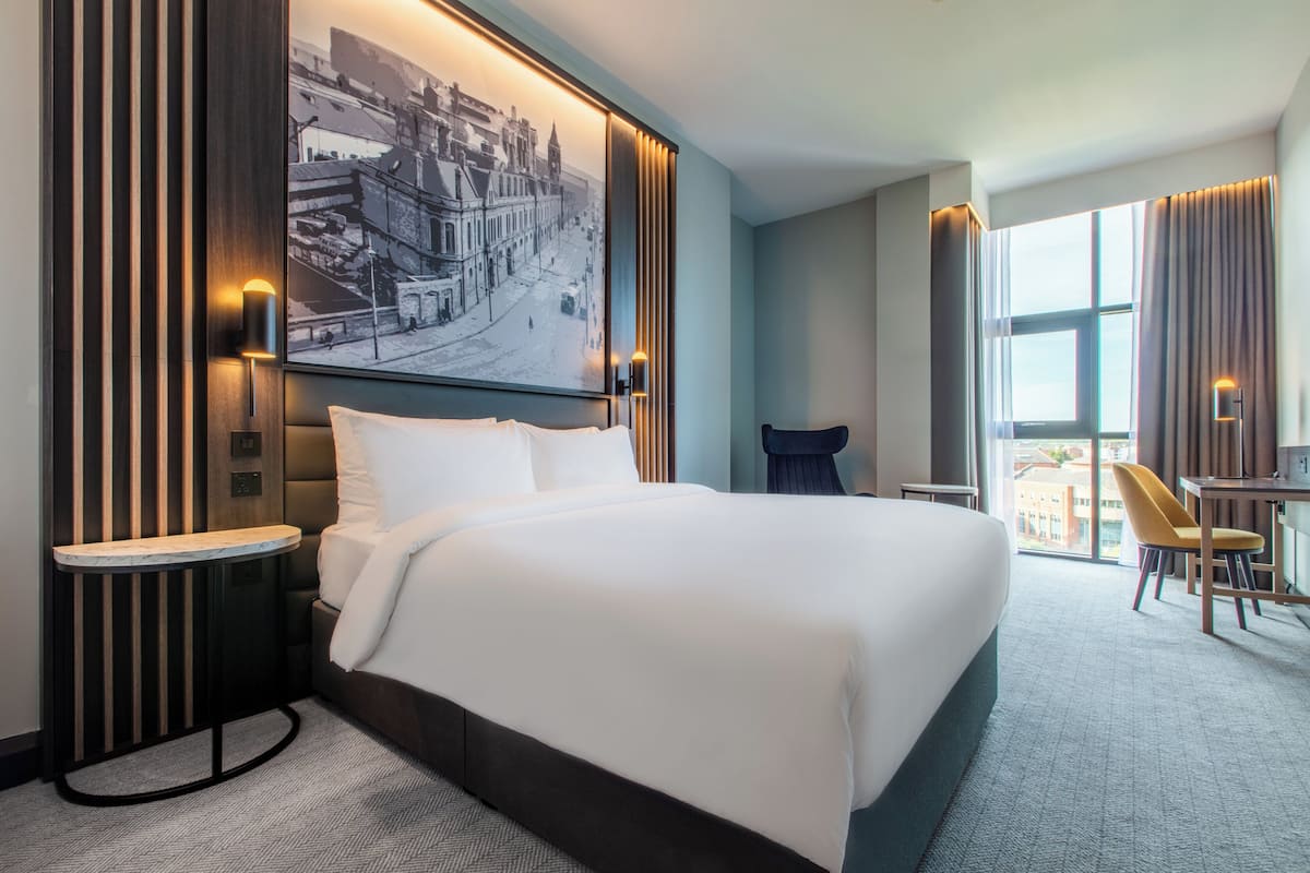 A modern hotel room with a large bed, a desk, a chair, and a large window.