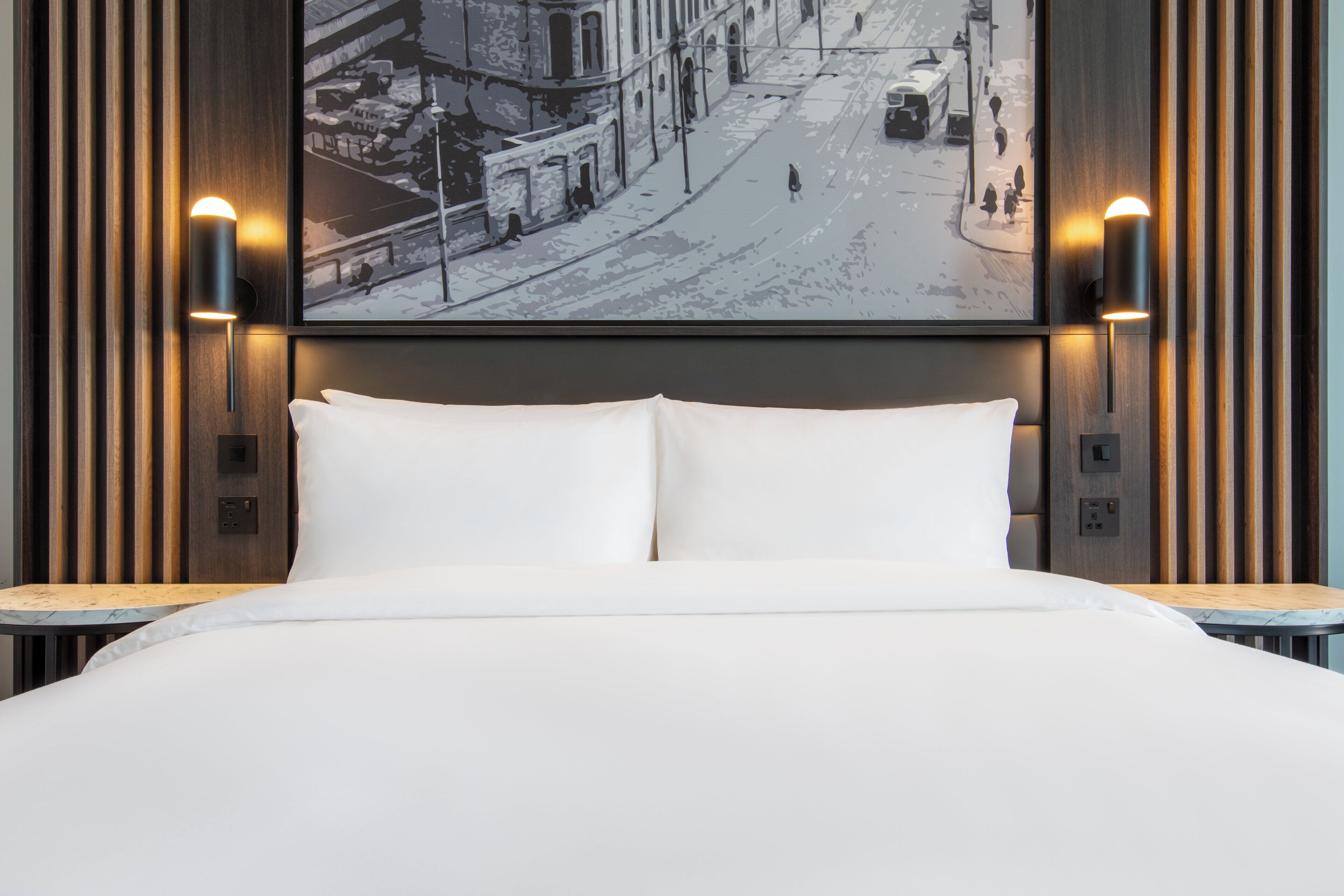A hotel room with a large bed, two wall-mounted lamps, and a cityscape painting on the wall.