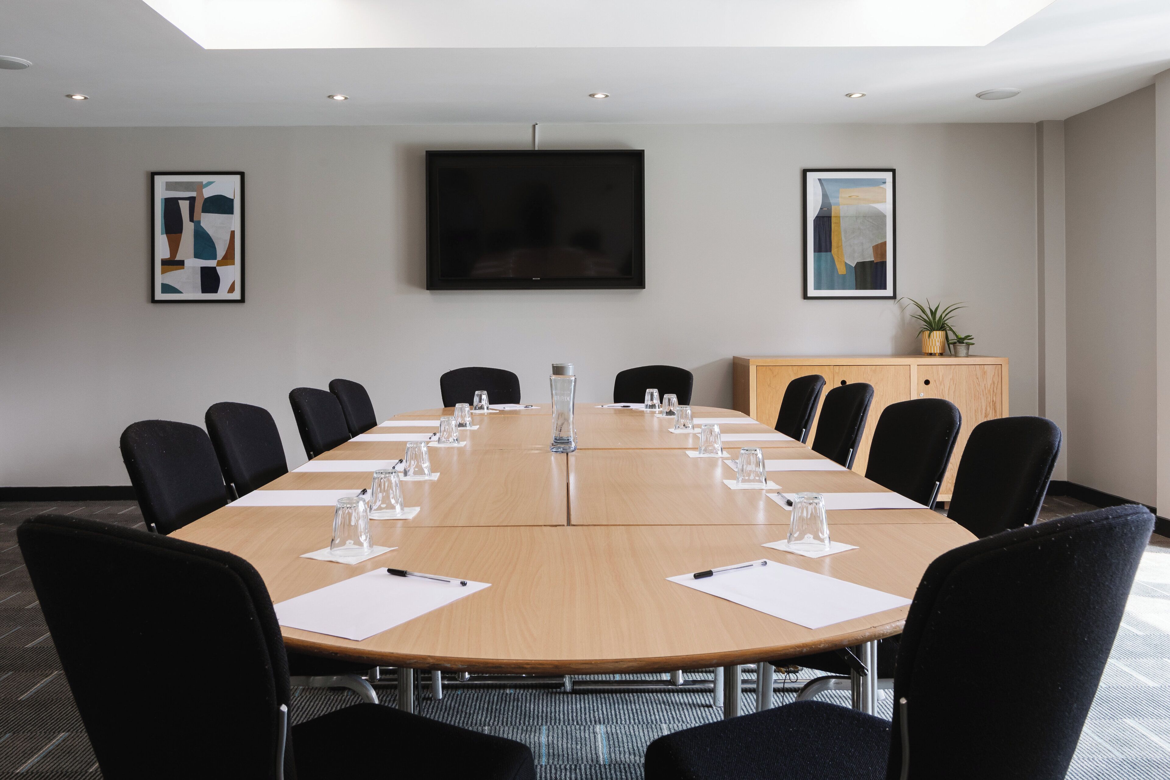 A conference room with a large table, black chairs, a TV, and artwork on the walls.